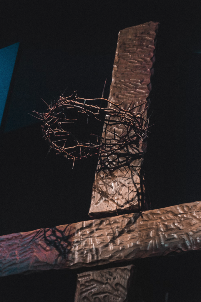 A textured wooden cross adorned with a crown of thorns, illuminated in a dimly lit setting with dramatic shadows.