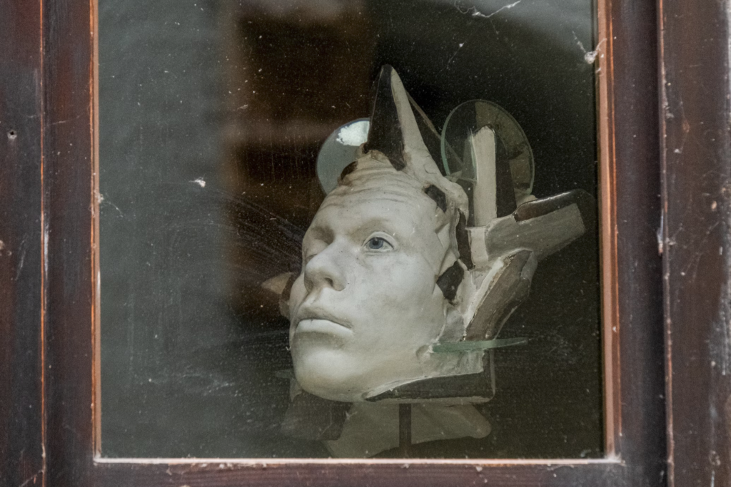 A fragmented sculpture of a human face with mechanical-like attachments, displayed behind a glass window in a wooden frame.