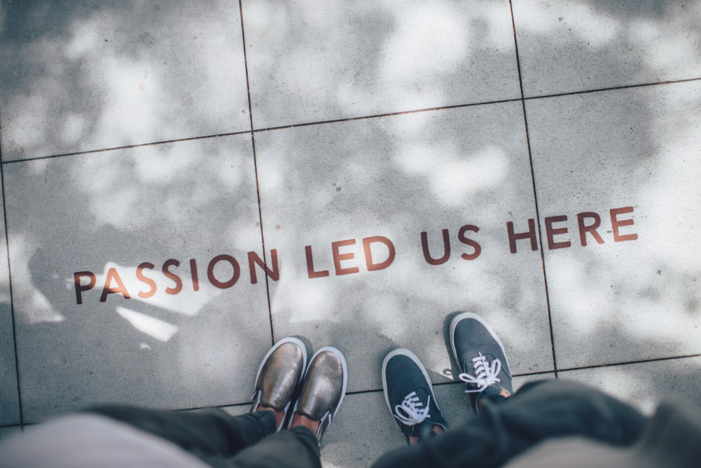 A pair of feet standing on a pavement with the words 'Passion Led Us Here' engraved on the tiles, surrounded by shadows.