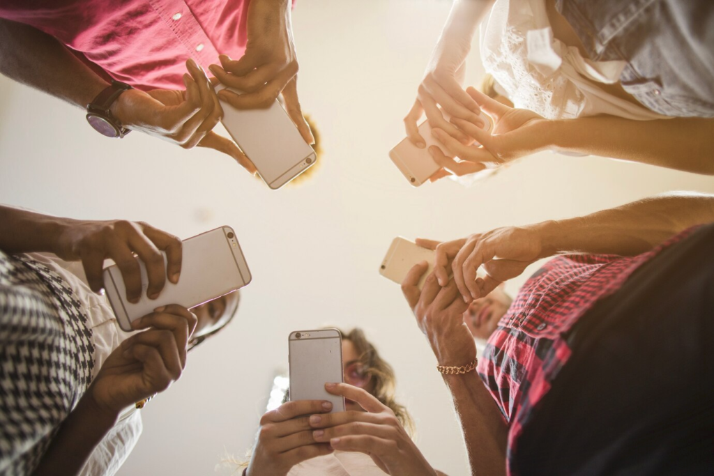 A diverse group of people standing in a circle, holding smartphones, captured from a low-angle perspective against a bright background.